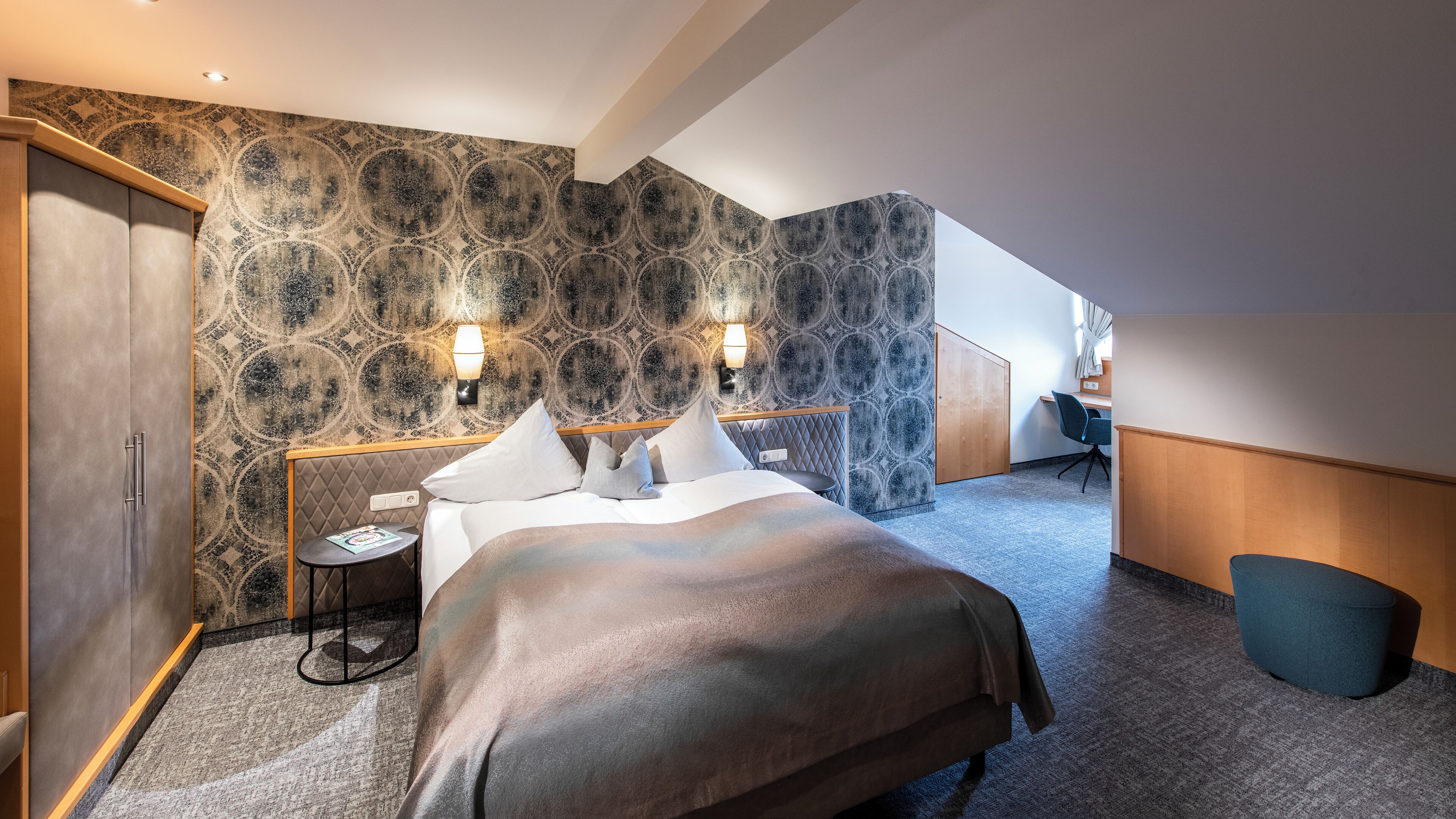 A modern hotel room with a large bed, two bedside lamps, a small table, and a desk with a chair.
