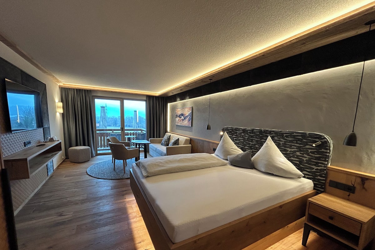 Habitación de hotel moderna con una cama grande, televisor montado en la pared, balcón con vistas y suelo de madera.
