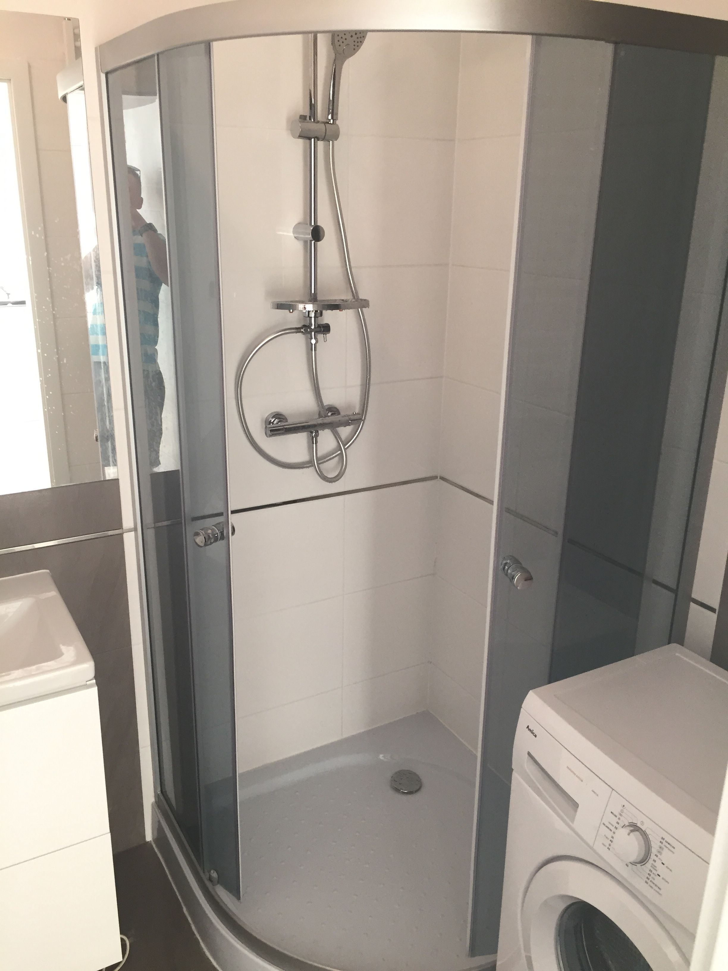 A small, enclosed shower area with a glass door, a washing machine, and a toilet in the adjacent space.