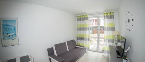 A modern living room with a sofa, a dining table with chairs, a small desk with a computer, and a window with curtains.