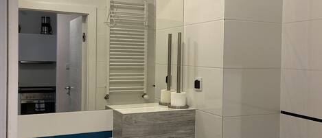 A modern bathroom with a white sink, mirror, and toilet. There is a towel rack above the sink and a heater on the wall.