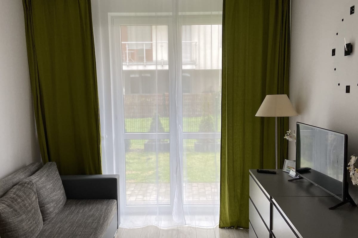 Un salon moderne avec un canapé gris, un petit bureau avec un téléviseur et une grande porte-fenêtre coulissante donnant sur un jardin.
