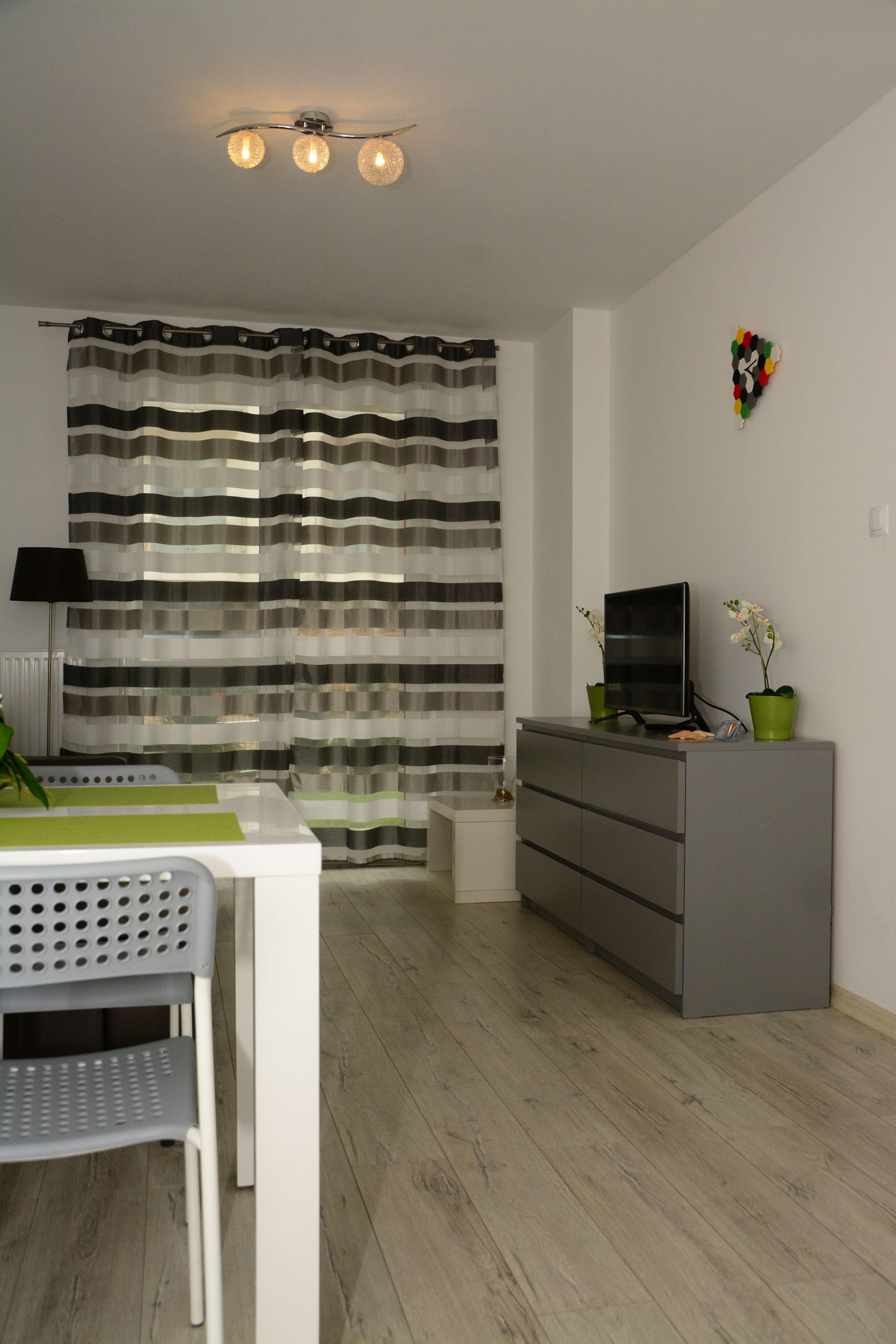 A modern living room with a striped curtain, a flat-screen TV on a grey cabinet, a small table with a green placemat, and a grey chair.