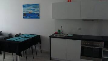 A compact kitchen with a dining area, featuring a black table, white cabinets, and a painting on the wall.