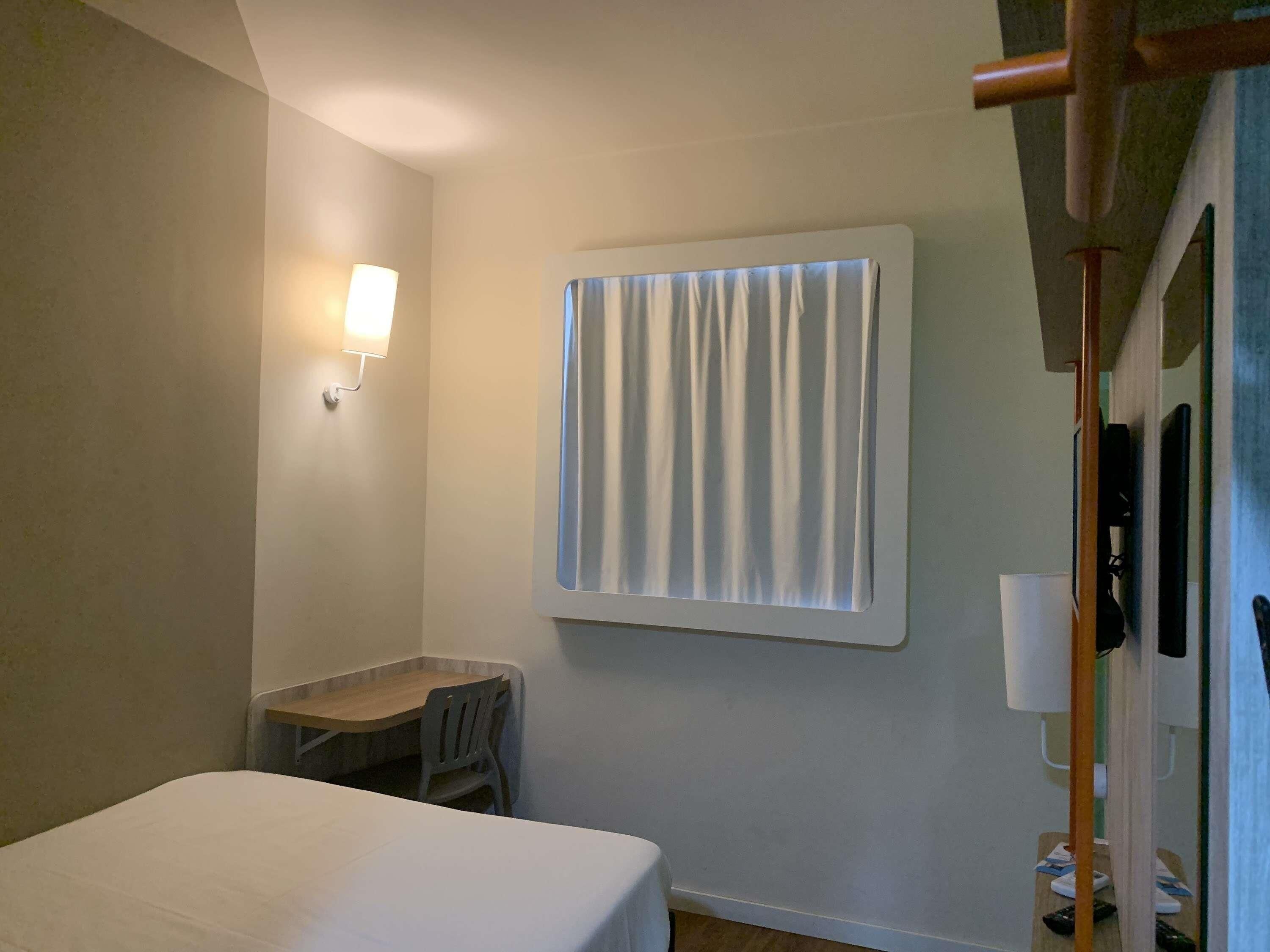 A small hotel room with a bed, a desk, a chair, a window with curtains, a wall-mounted lamp, and a wooden door.