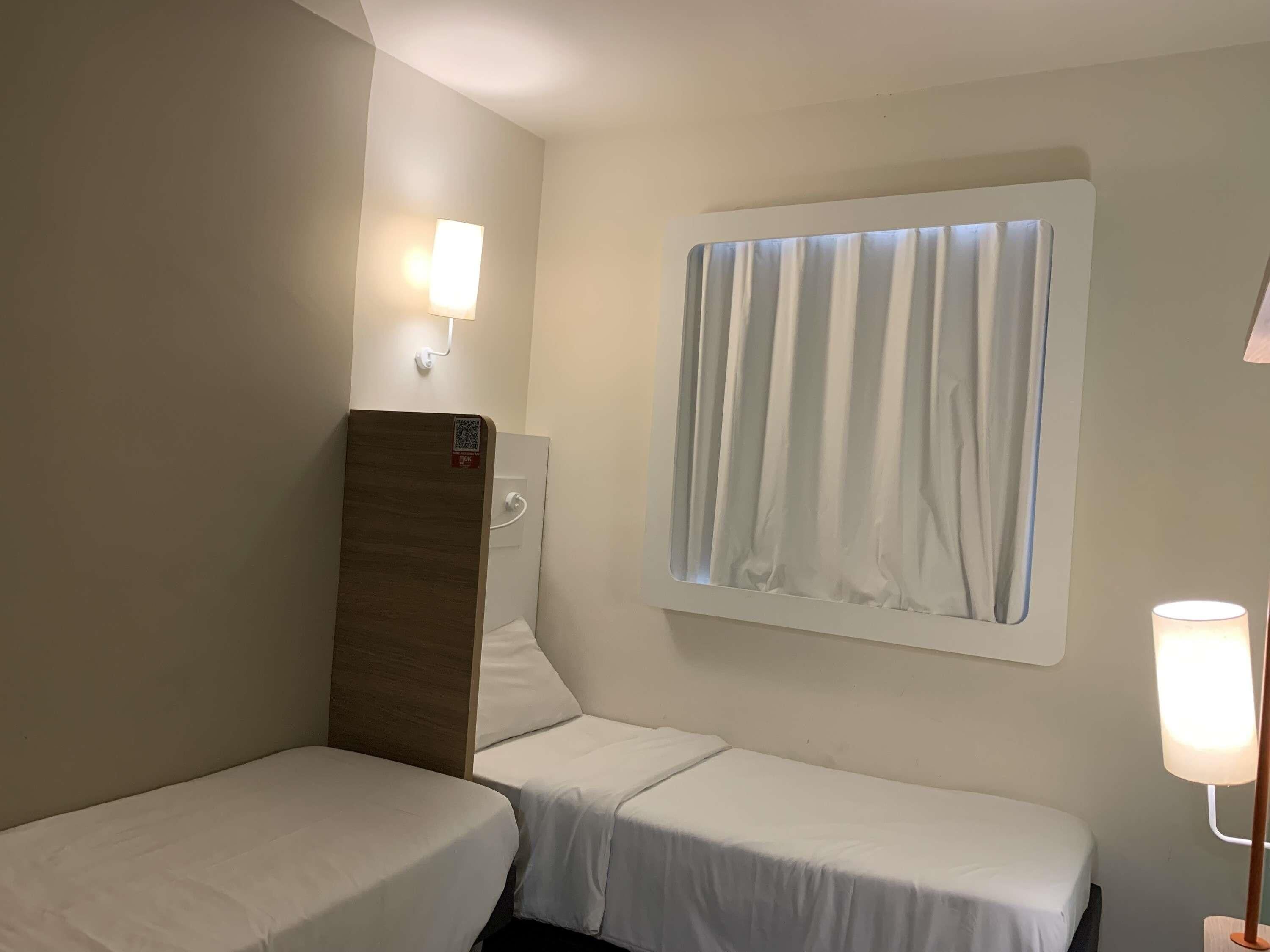 Et hotelværelse med to enkeltsenge, et vindue med gardiner fastgjort på væggen, et natlys og en QR-kode på sengegavl.