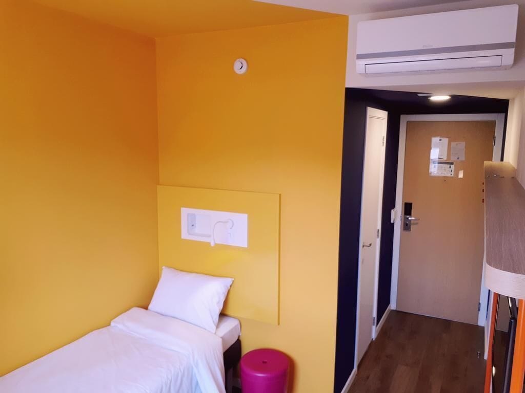 Una piccola camera con un letto singolo, una parete gialla, un letto bianco e un cestino rosa, con una porta che dà su un'altra stanza.