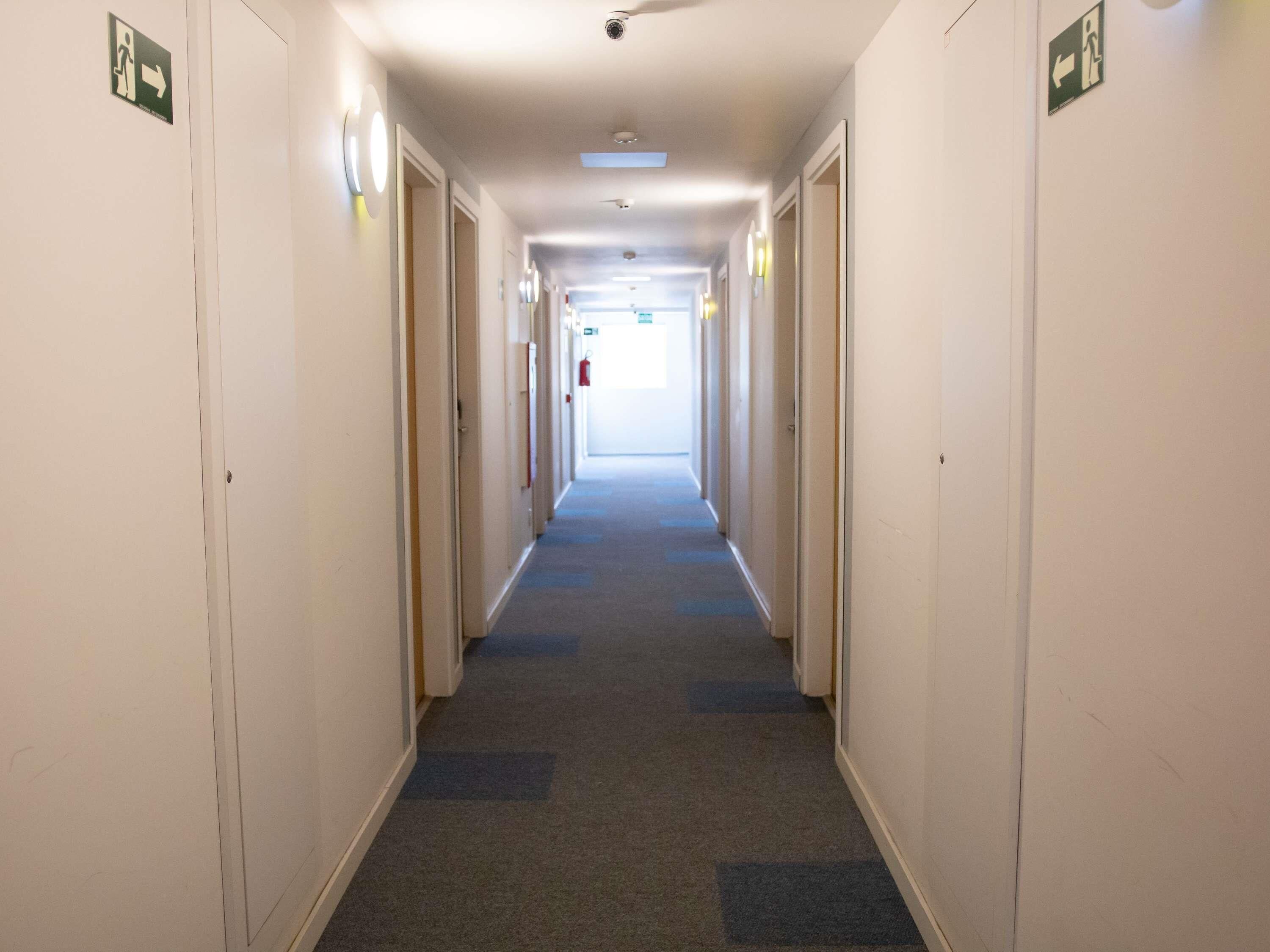 Um corredor de hotel comprido e estreito, com portas em ambos os lados, piso coberto de carpete e placas de saída de emergência.