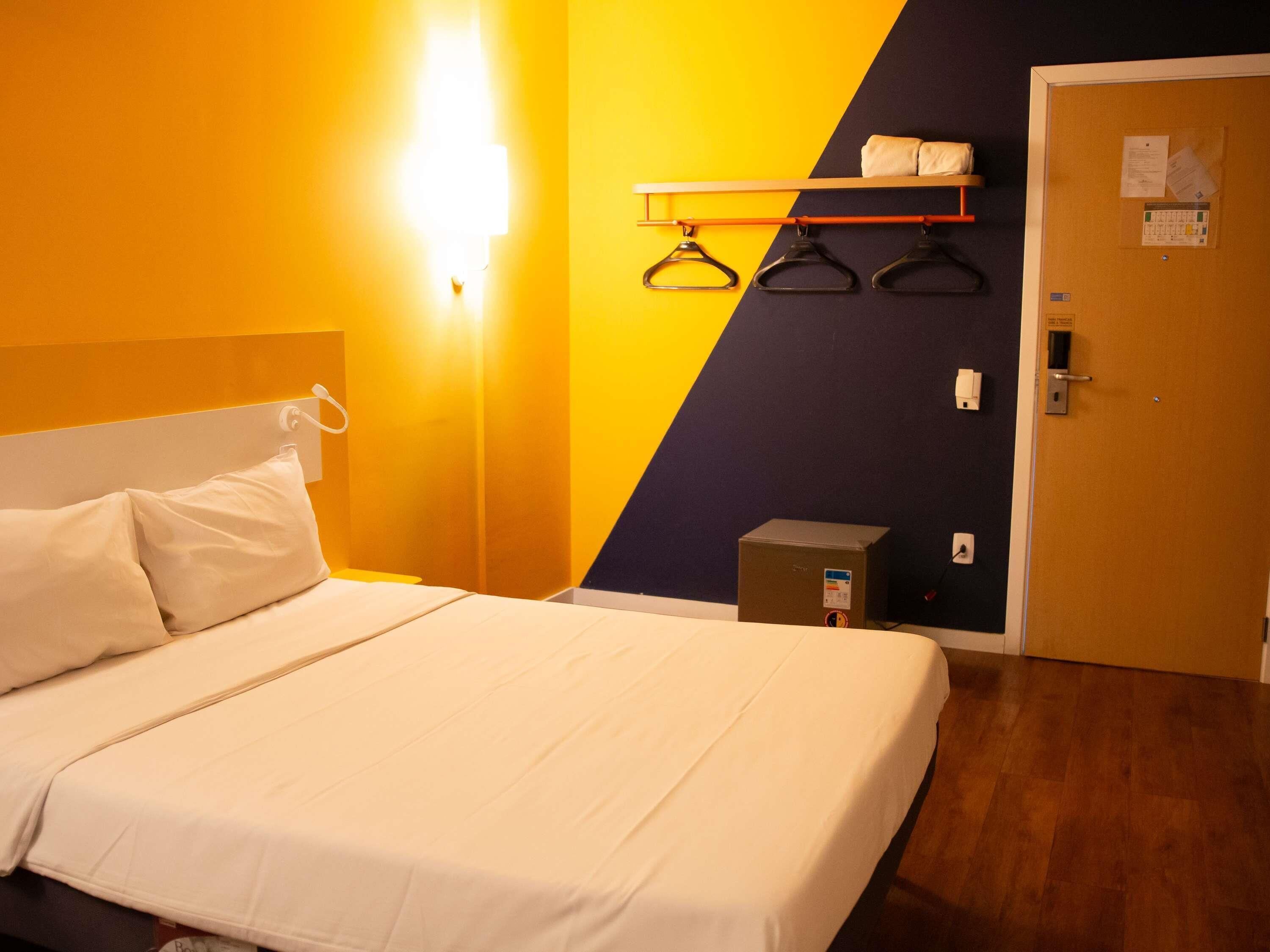 Une chambre d’hôtel avec un lit, une lampe fixée au mur, un porte-manteau, une porte et une petite boîte posée sur le sol.