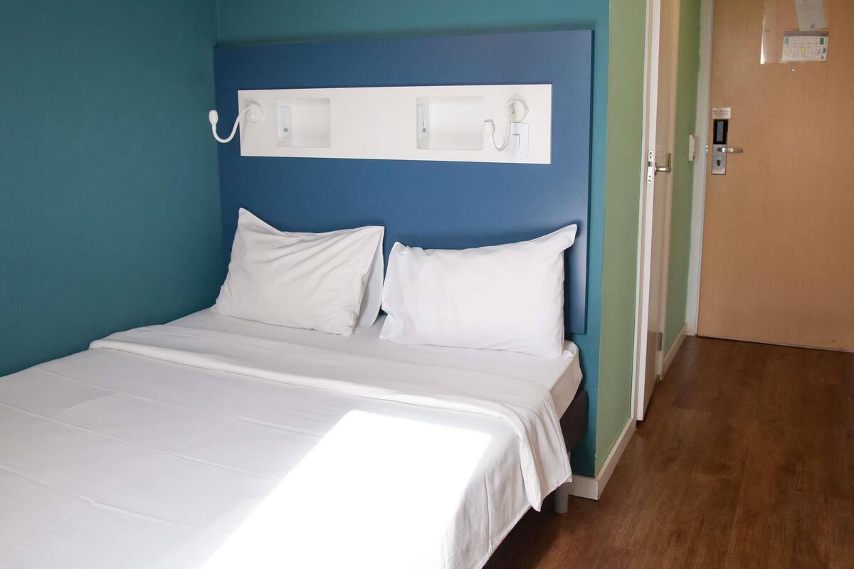 Habitación de hotel con cama, ropa de cama blanca, cabecero azul y piso de madera.