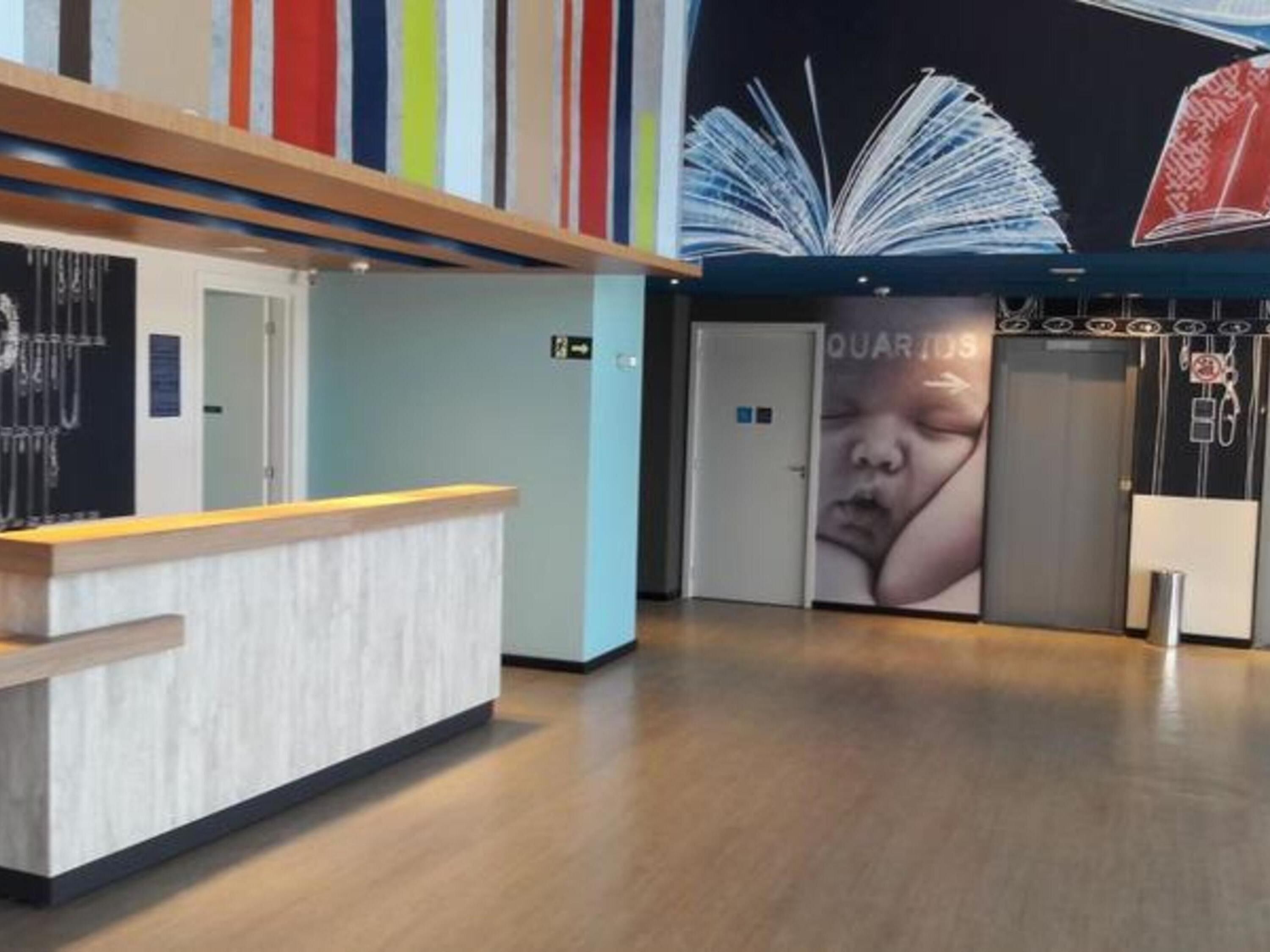 A modern reception area with a wooden reception desk, a large mural of books on the wall, and a visible elevator door labeled "QUARTEES".
