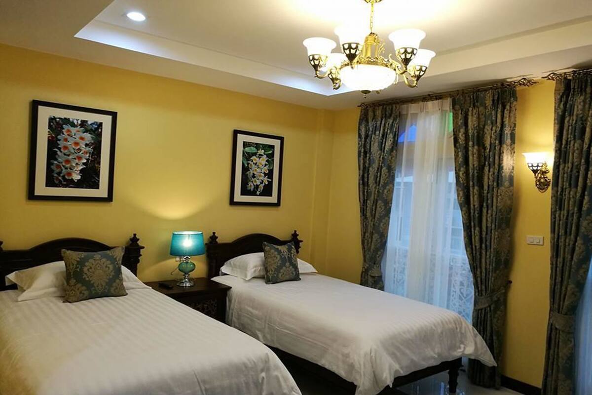 A hotel room with two beds, a chandelier, framed floral artwork on the walls, and a window with curtains.