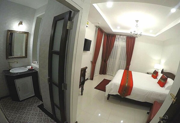 Habitación de hotel con cama, mesita de noche, lámpara, ventana con cortinas, espejo y baño con lavabo e inodoro.