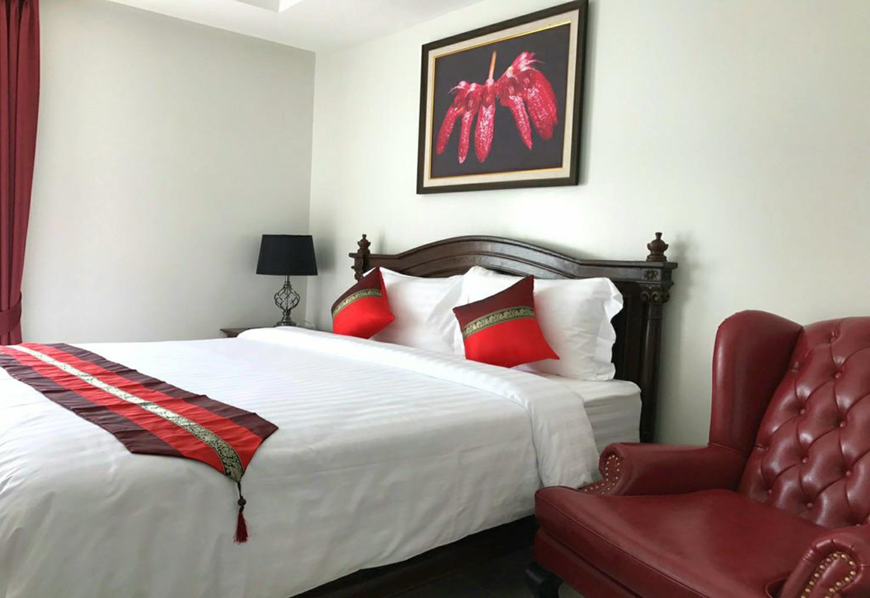 A neatly made bed with white linens and red accents, a red leather armchair, and a framed picture on the wall.