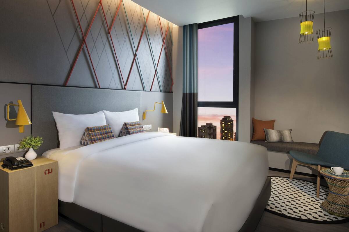 A modern hotel room with a large bed, a city view, a small seating area, and a unique geometric wall design.