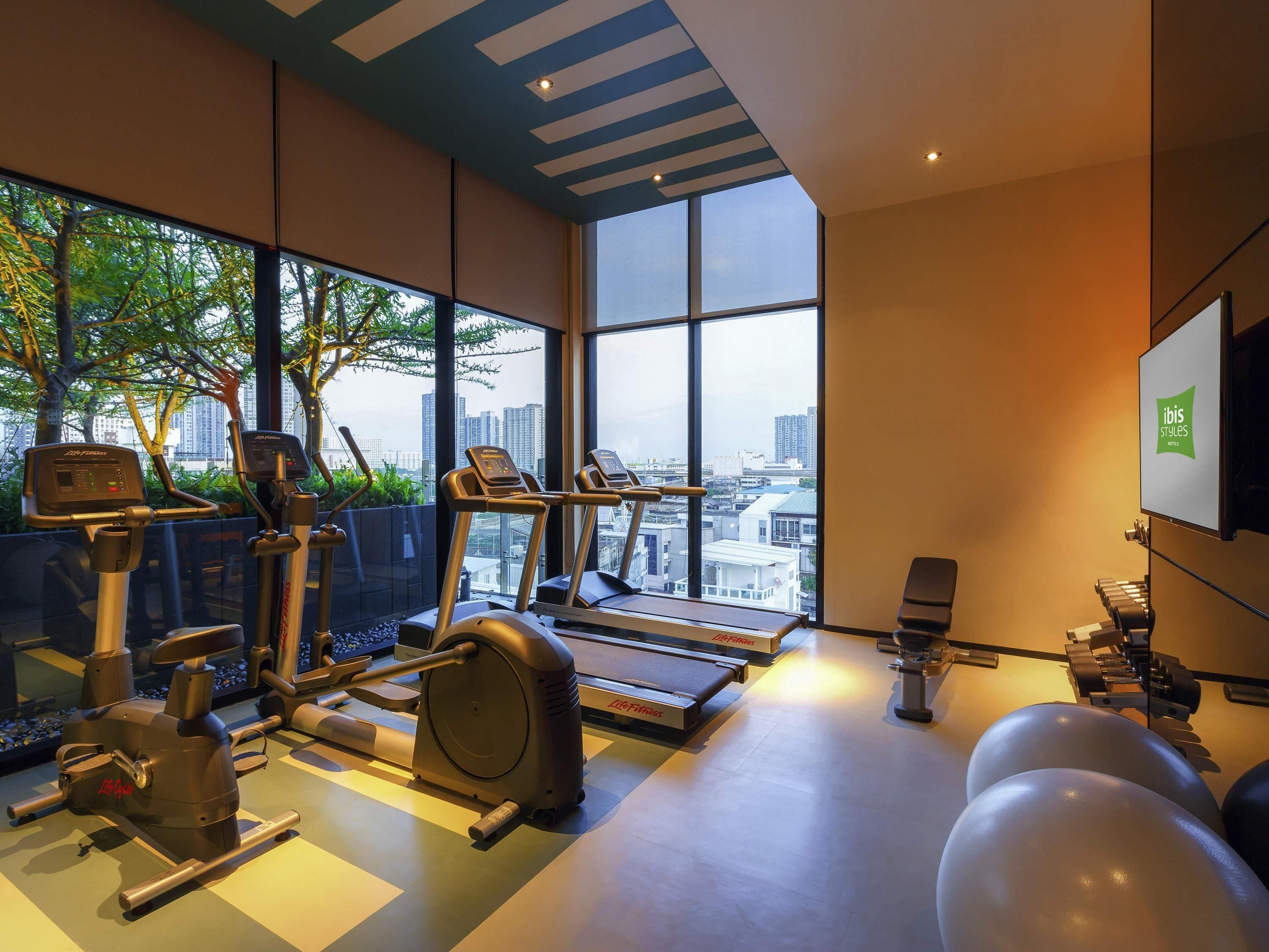 Ett gym med träningsutrustning, ett löpband och ett stort fönster med utsikt över staden.