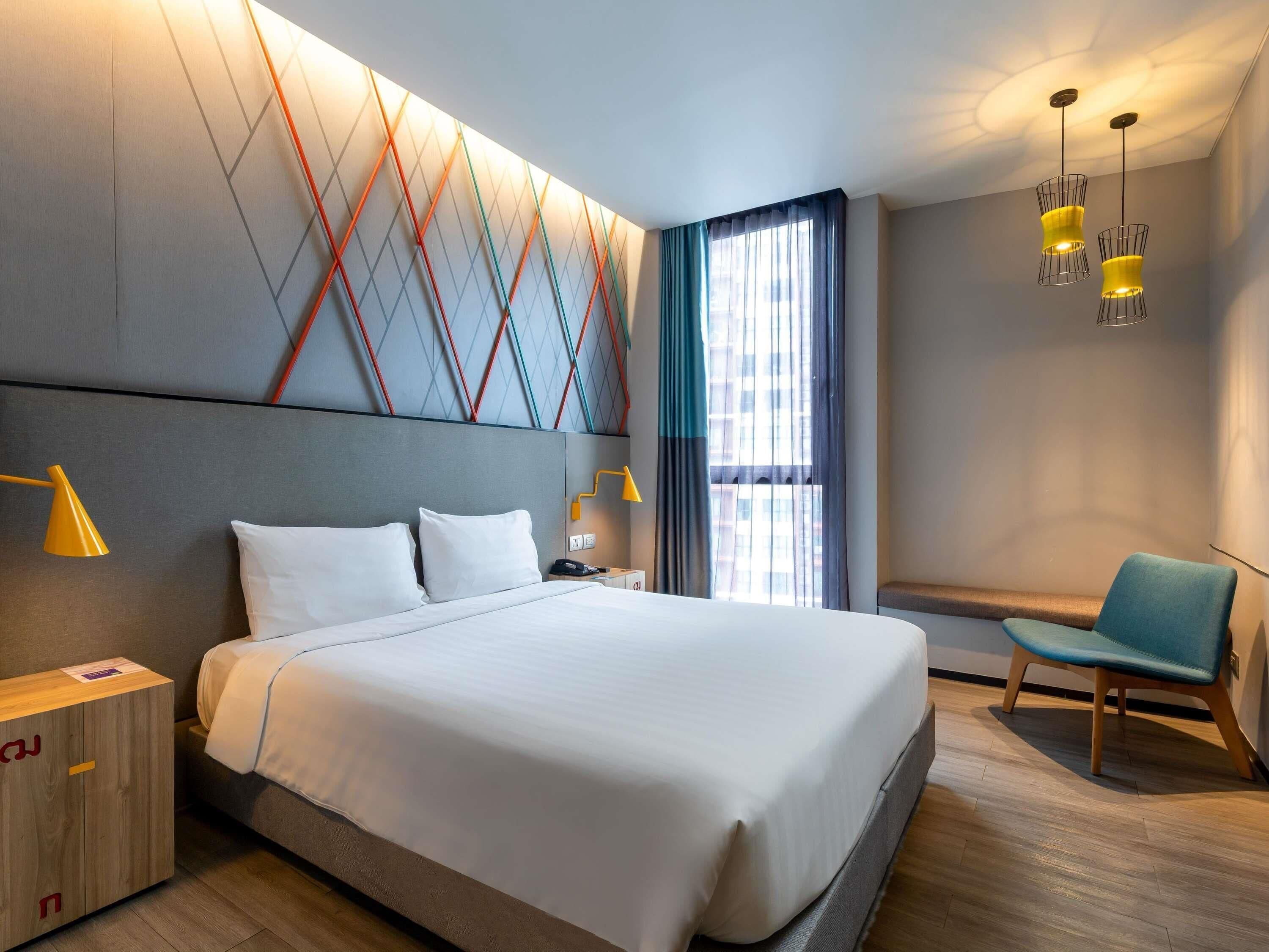 A modern hotel room with a large bed, bedside lamps, a seating area, and a view of the city through the window.
