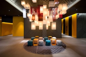 Um lobby moderno com padrões geométricos nas paredes, uma área de estar central com pufes coloridos e luminárias pendentes.