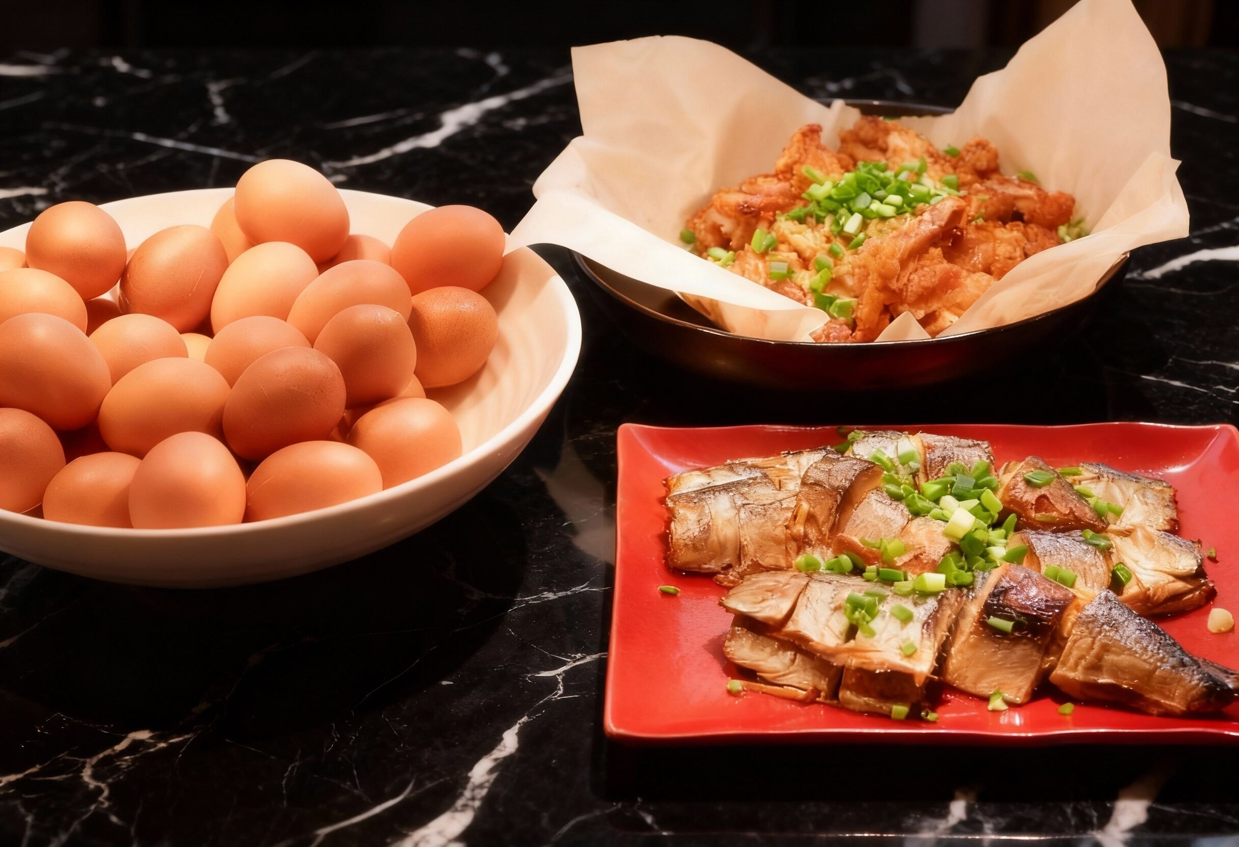 A marble countertop with three dishes: a bowl of eggs, a pan of fried chicken, and a plate of sliced meat with green onions.