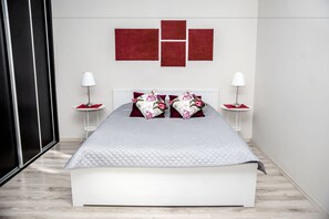 Ein modernes Schlafzimmer mit einem weißen Bett, roten und floralen Kissen sowie zwei Nachttischen mit Lampen.