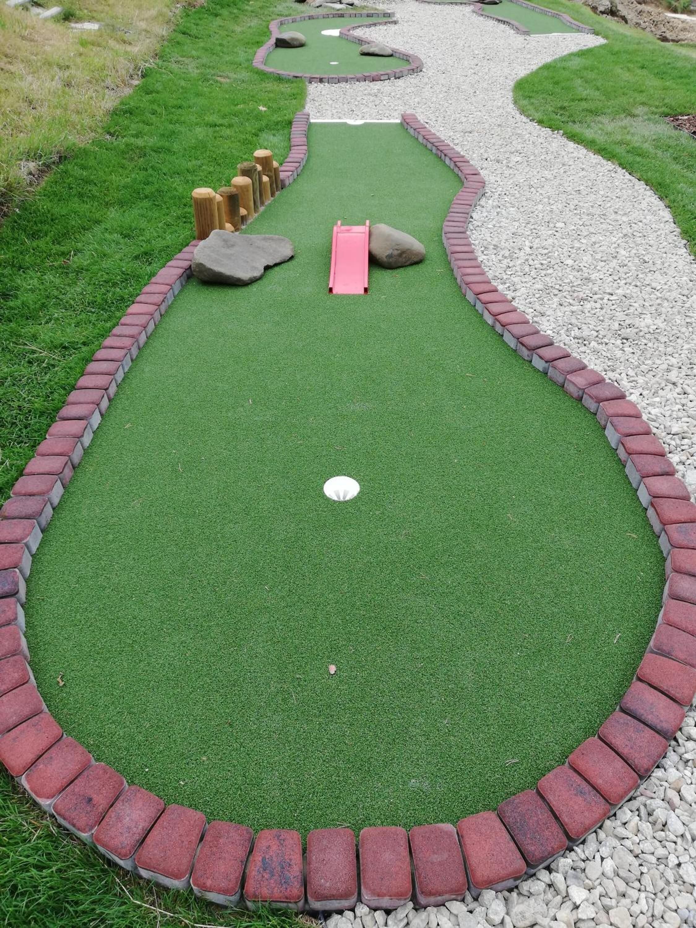 Un campo de mini golf con césped artificial verde, un borde de ladrillos rojo y blanco, y un hoyo para pelotas blancas.