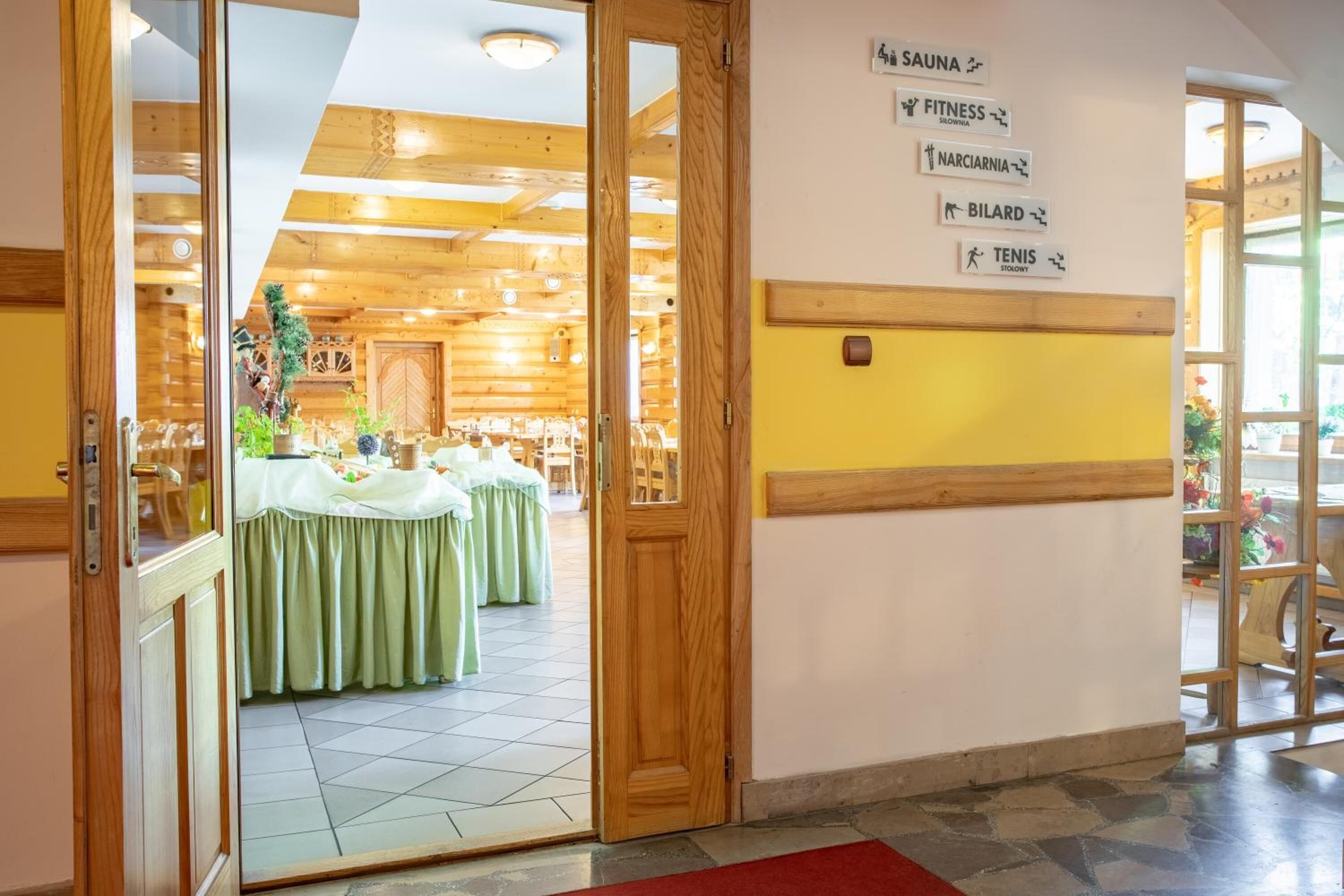 Un vestíbulo de hotel con interior de madera, una mesa de recepción y una pared con indicaciones hacia diversas instalaciones como sauna, gimnasio y tenis.