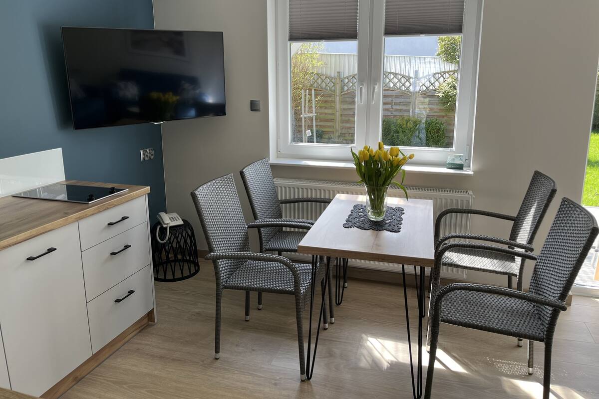 A modern kitchen with a flat-screen TV, a dining table with chairs, and a window with blinds.