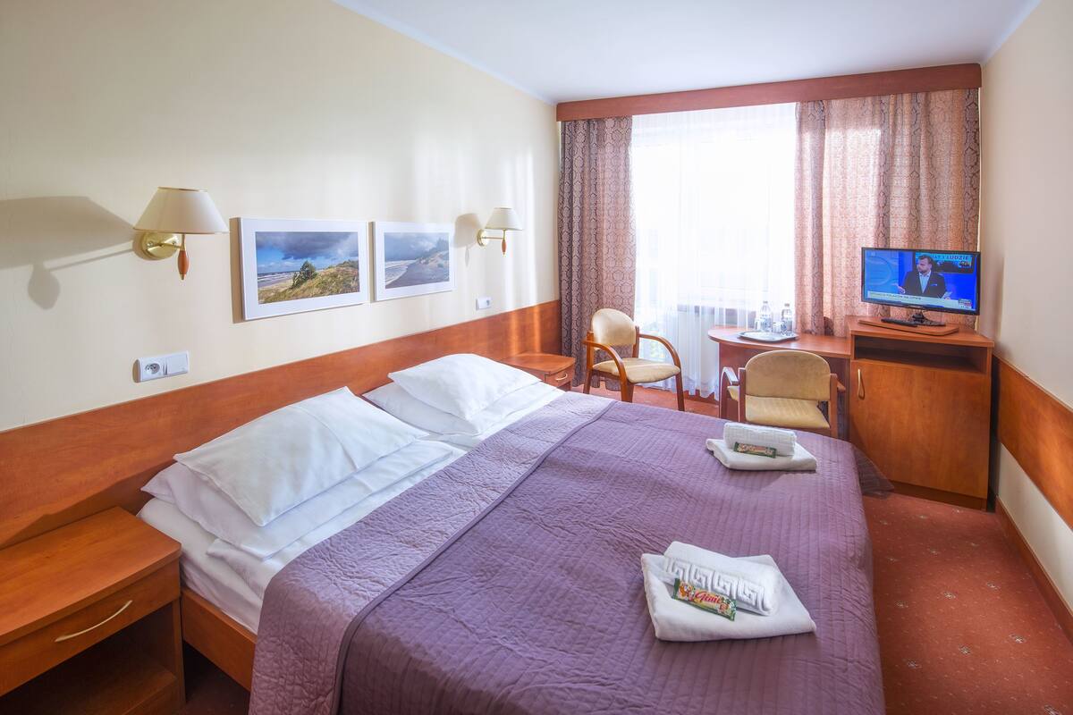 A hotel room with a large bed, a desk with a chair, a television, and two wall-mounted lamps.