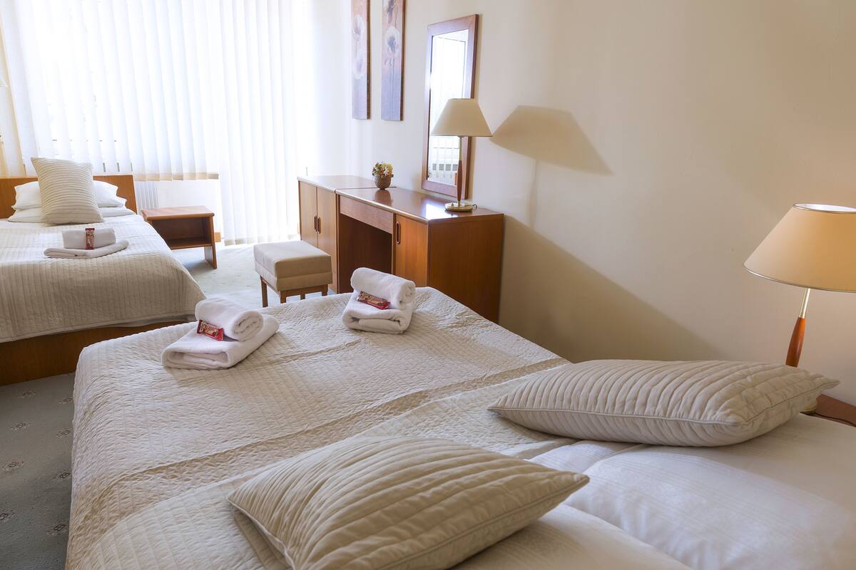 A hotel room with two beds, a desk, a chair, a lamp, and a window with blinds.