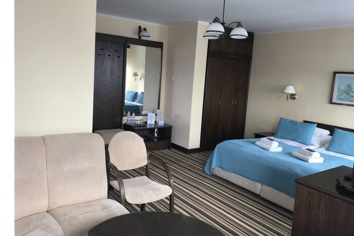 A hotel room with a bed, a sofa, a chair, a desk, a mirror, and a ceiling light fixture.