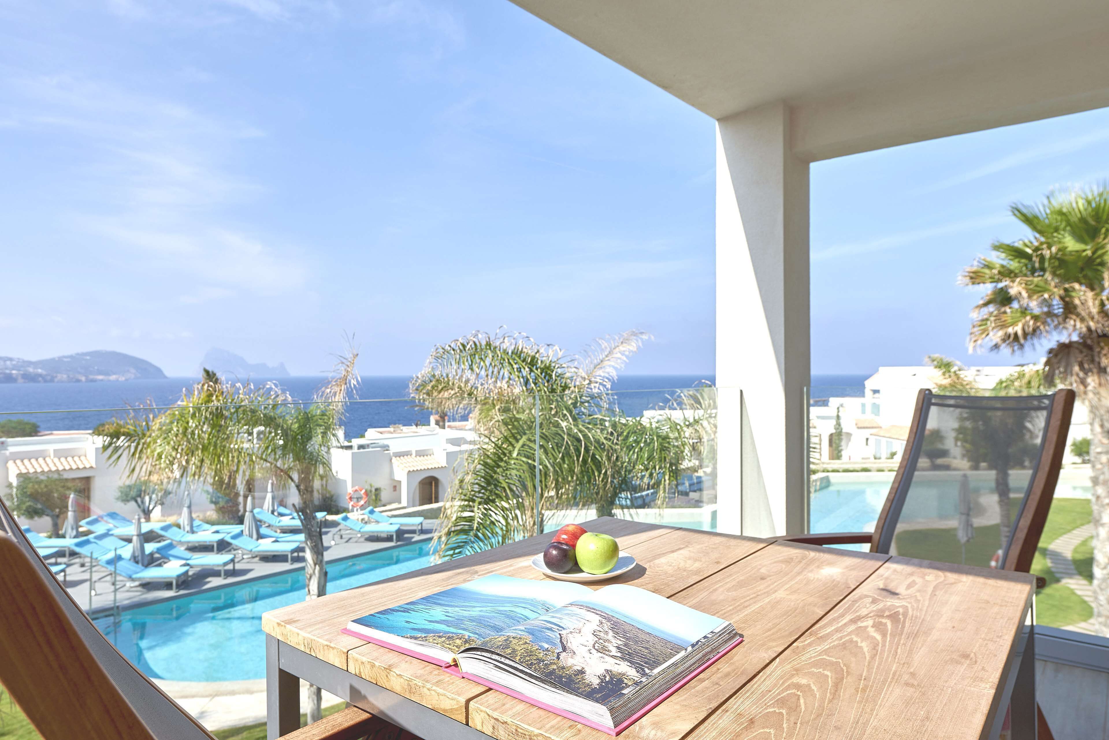 Une table en bois, sur laquelle se trouvent un livre et un saladier rempli de fruits, donnant sur une piscine et offrant une vue sur la mer.