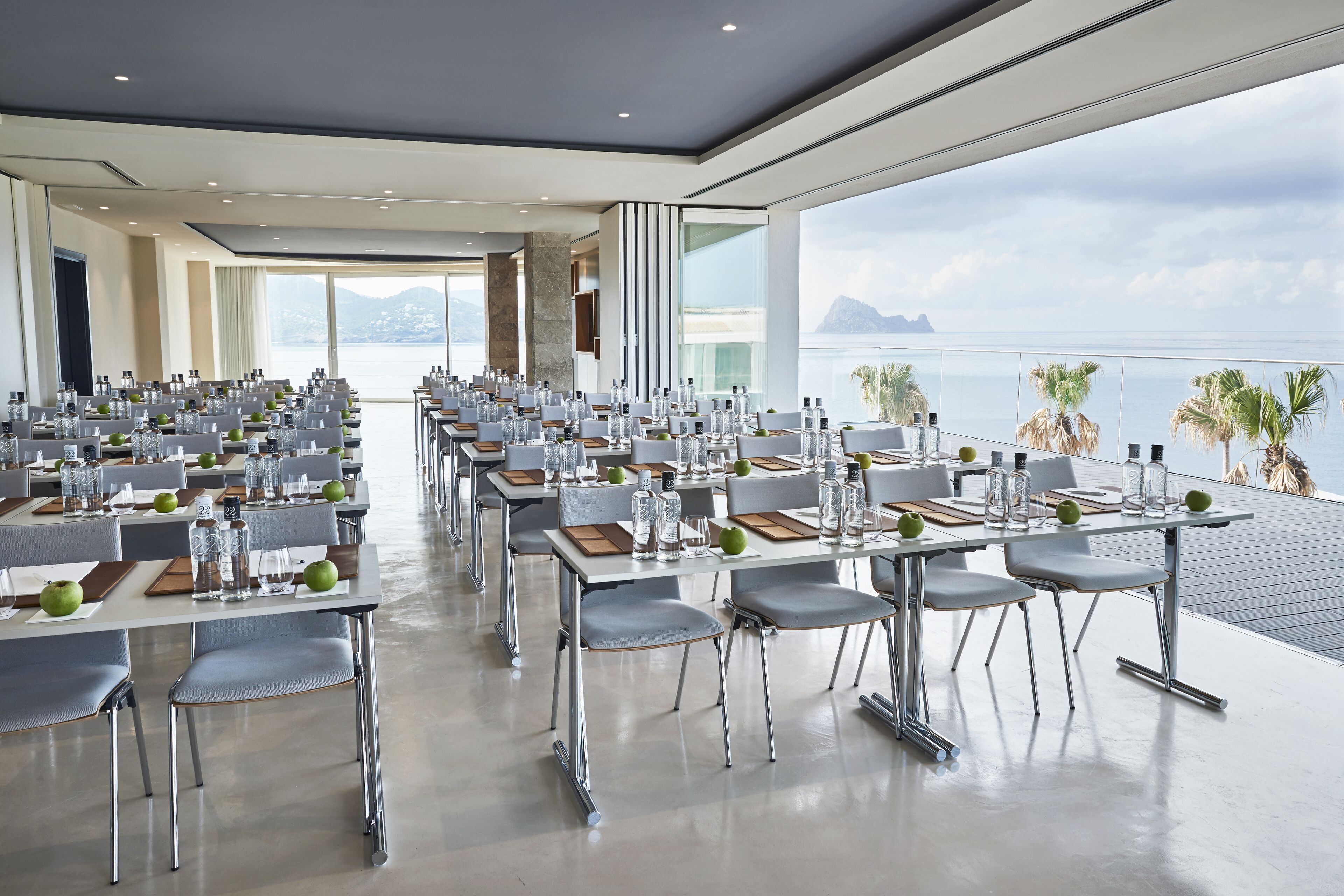 Un salón de conferencias con mesas dispuestas para una reunión, ventanas amplias con vistas al mar y a las montañas, y una decoración de diseño moderno.