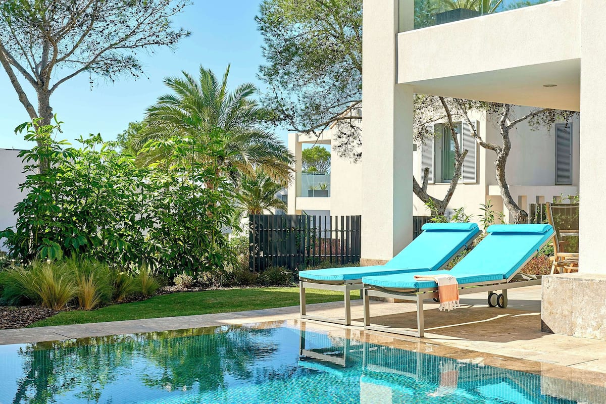 Una piscina con dos sillones de descanso, rodeada de vegetación y un edificio moderno.
