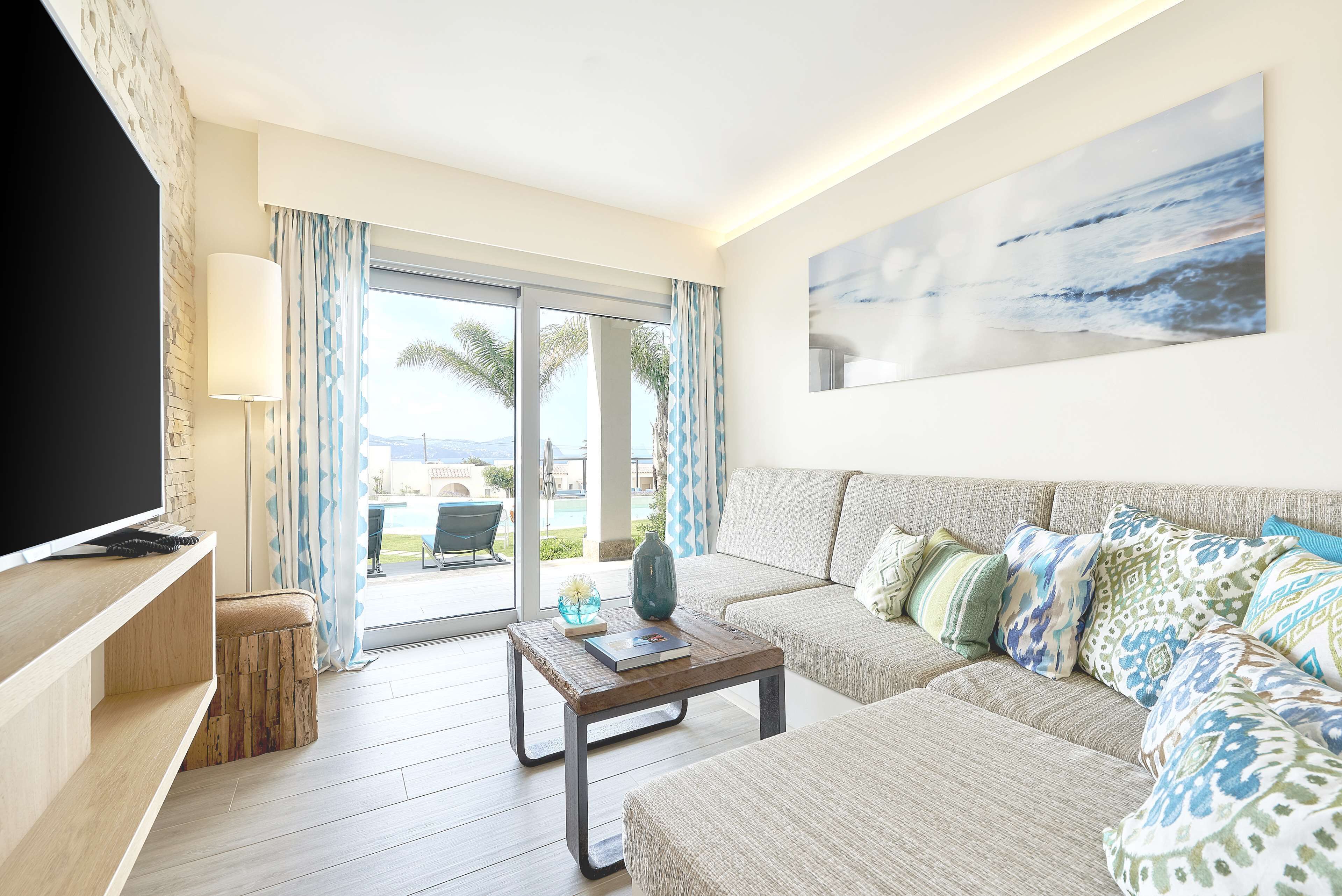 A modern living room with a sofa, a coffee table, and a TV. There is a view of palm trees and a pool through the glass doors.