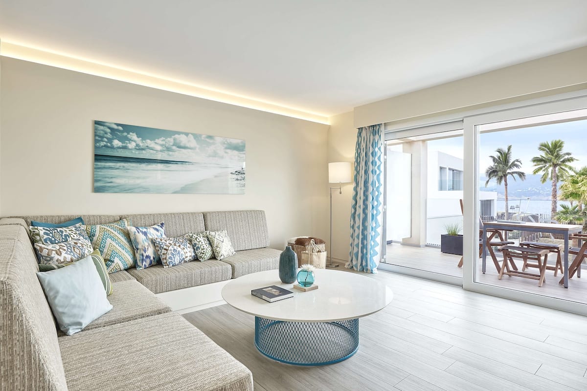 Una sala de estar moderna con un sofá, una mesa redonda de centro y un ventanal con vistas a palmeras y a la playa.