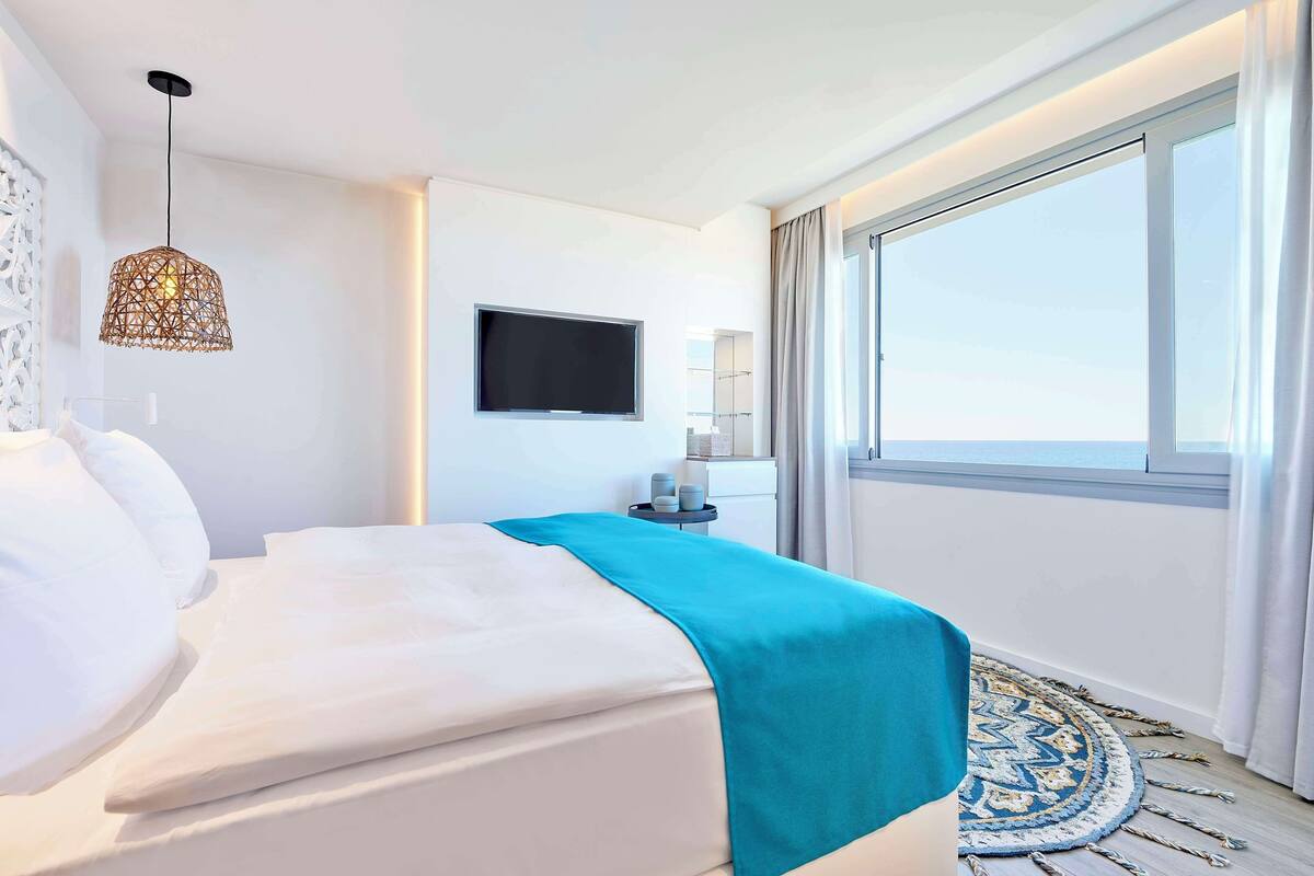 Una habitación de hotel moderna con una cama grande, un televisor montado en la pared y un ventanal con cortinas.