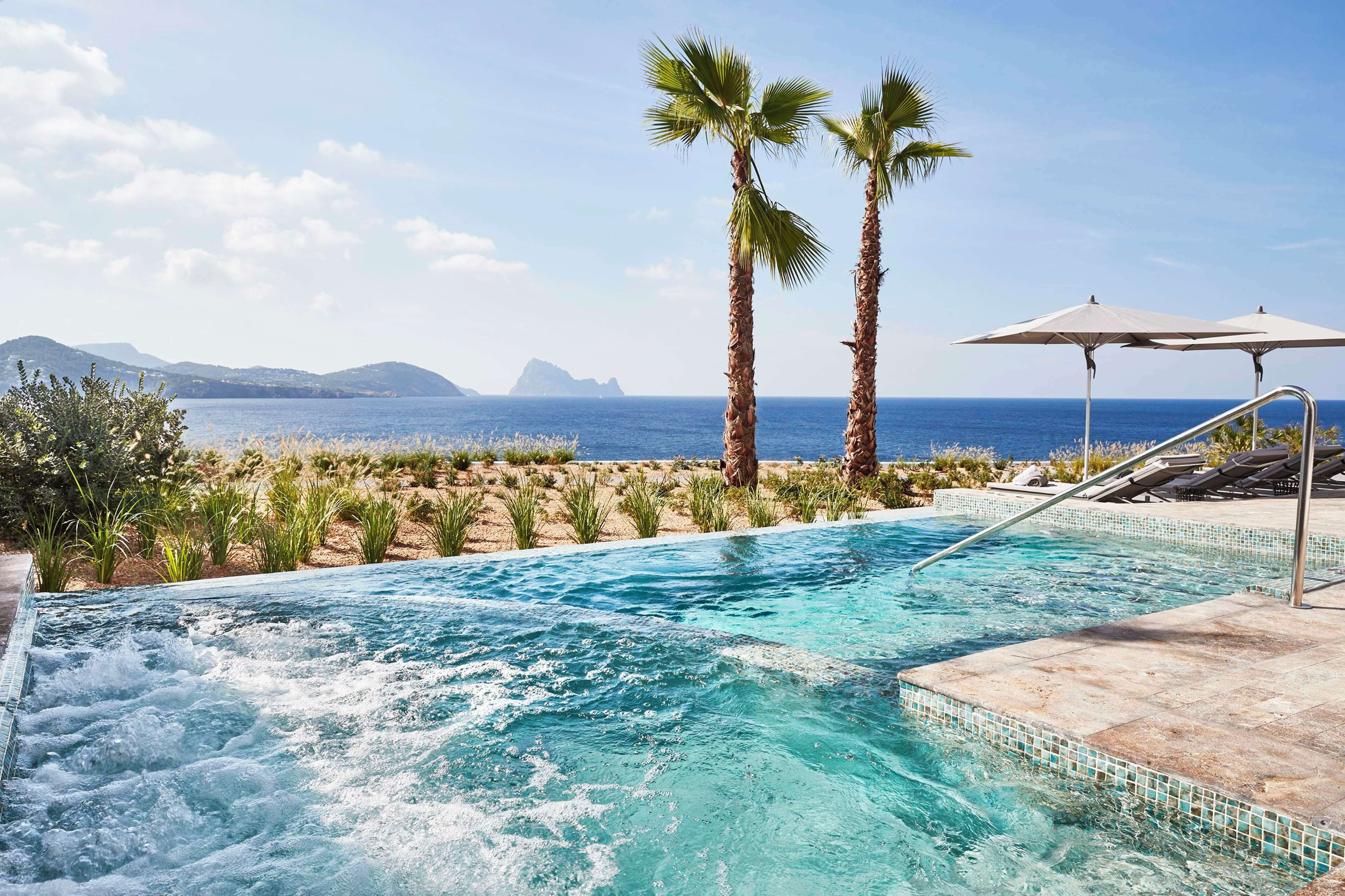 Una piscina con vistas al mar y a las montañas, rodeada de palmeras y sillas de descanso.