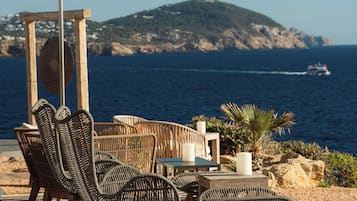 Outdoor seating with wicker chairs and a table, overlooking a body of water and a mountainous landscape.