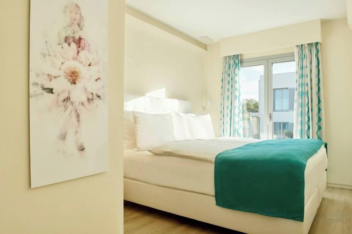 A bedroom with a bed, a window with curtains, and a painting on the wall.