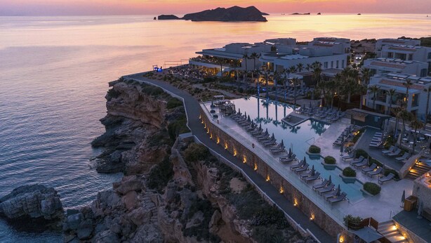 Un resort costero con zona de piscina, sillones de descanso y vistas al mar al atardecer.