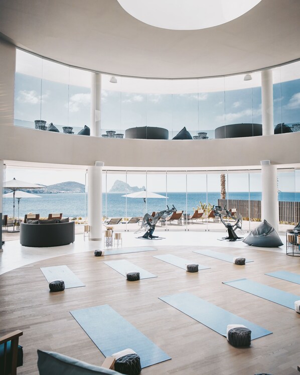 Amplio espacio interior con grandes ventanales con vistas al mar, que incluye colchonetas de yoga y equipo de ejercicio.