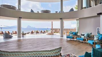 Un espacio interior amplio con suelo de madera, varios arreglos de asientos y ventanas grandes que ofrecen vistas al mar y a las montañas.