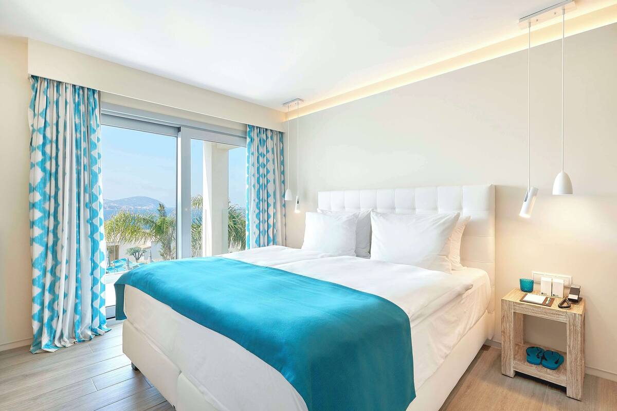 Habitación de hotel moderna con una cama grande, una mesita de noche con lámpara y vistas al exterior.