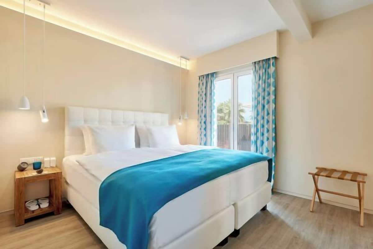 Una habitación de hotel moderna con una cama grande, una mesita de noche, un pequeño banco de madera y una ventana con cortinas.