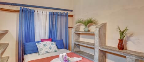 A hotel room with a bed, blue and white curtains, a wooden shelf with a plant, and a decorative towel on the bed.