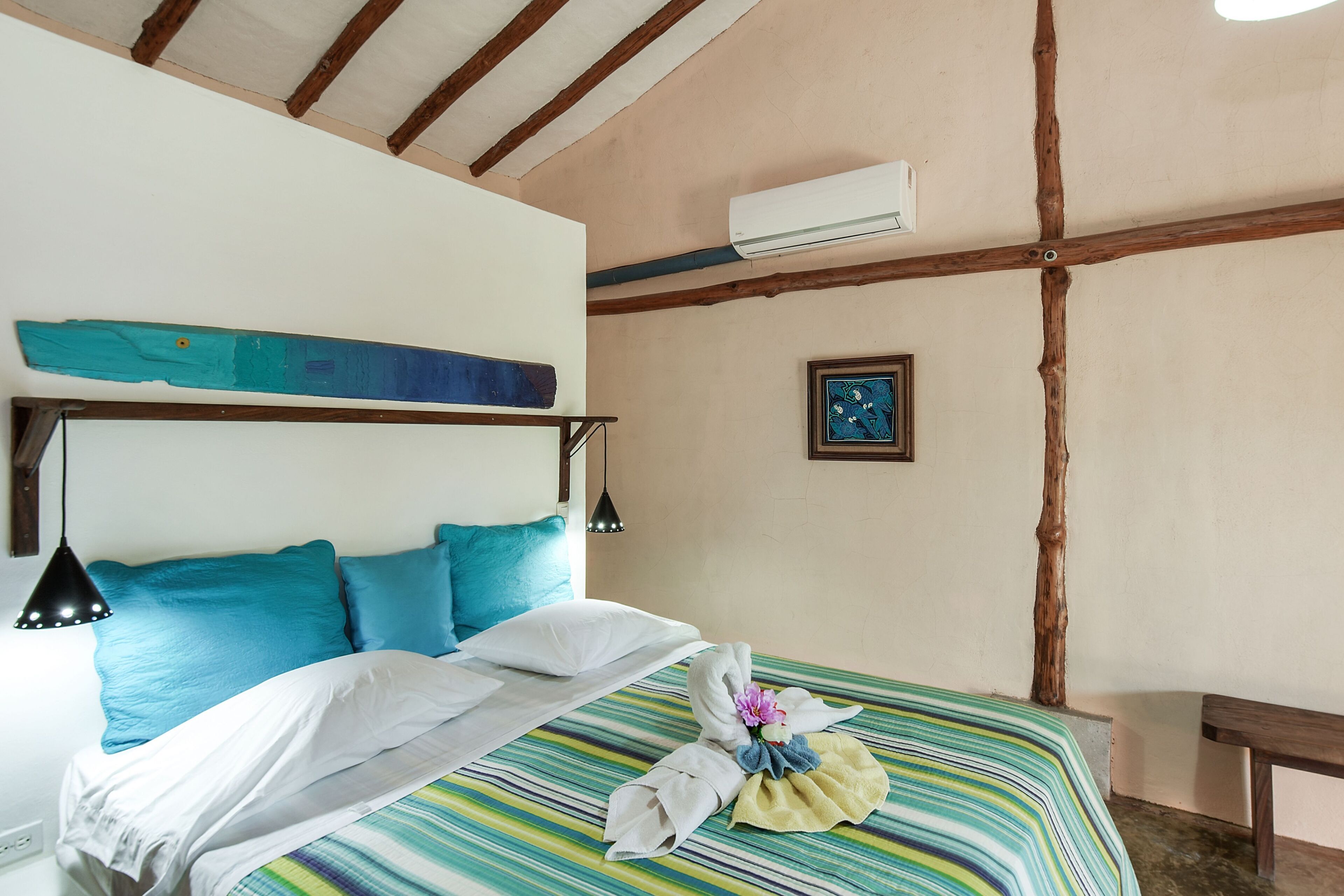 Een hotelkamer met een bed, hoofdbord, een wandgemonteerde airconditioner, een ingelijste foto en een houten bankje.
