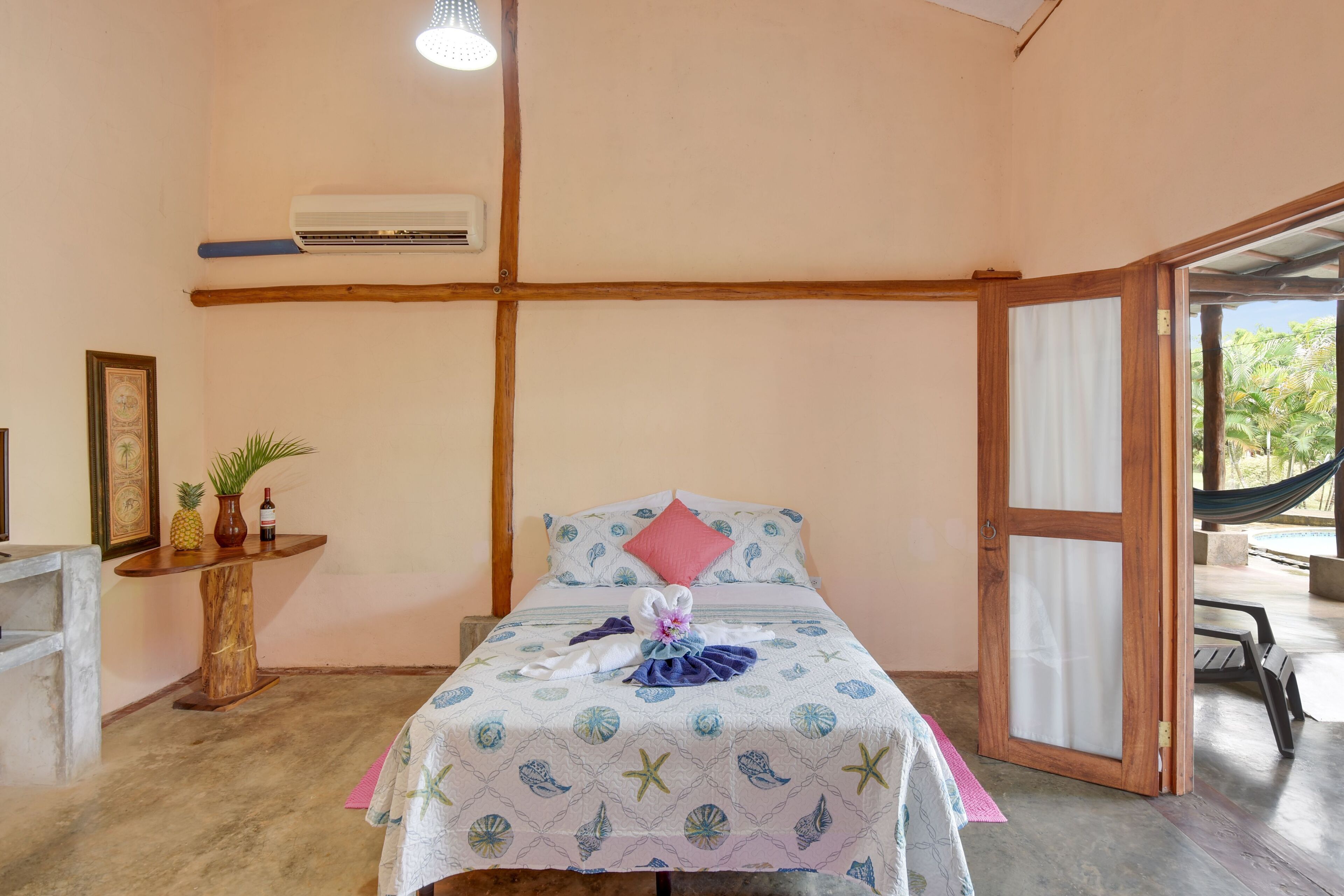 Une chambre avec un lit, une petite table sur laquelle est posée une plante, un tableau encadré et une porte donnant sur un balcon équipé d’un hamac.