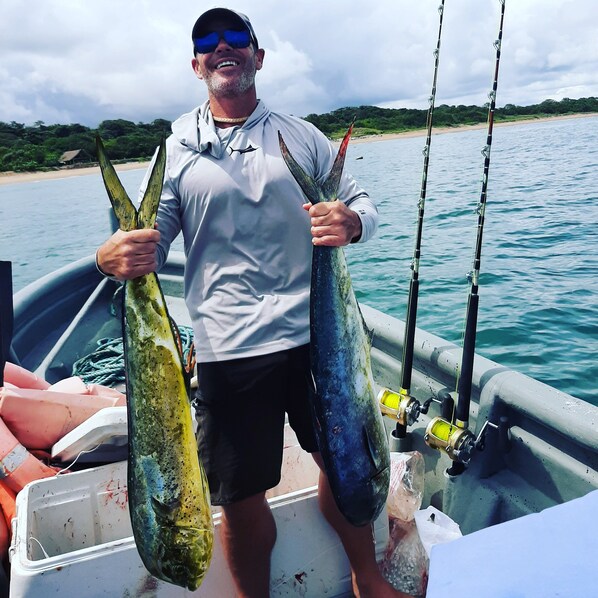A man holding two large fish on a boat with a beach and water in the background.