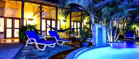 Un área de piscina nocturna con sillones reclinables y un edificio iluminado al fondo.