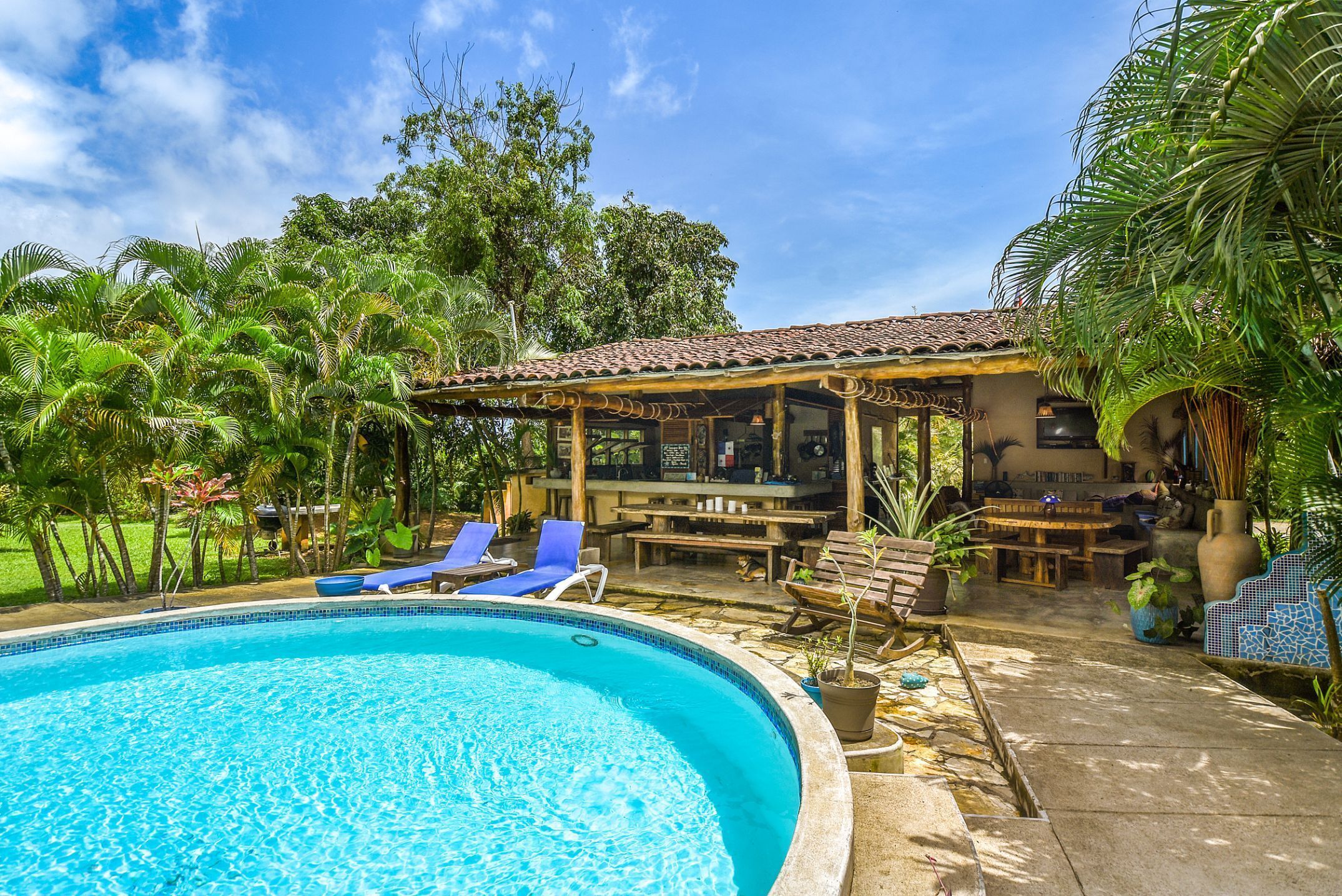 A swimming pool with lounge chairs, a covered outdoor seating area, and lush tropical vegetation.